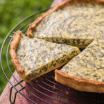 Klassische Spinat-Quiche in Nahaufnahme aus niedrigem Winkel auf einem schwarzen Kuchengitter, ein Stück angeschnitten und leicht abgehoben, mit goldenem Mürbeteigrand und grün gesprenkeltem Eier-Sahne-Guss