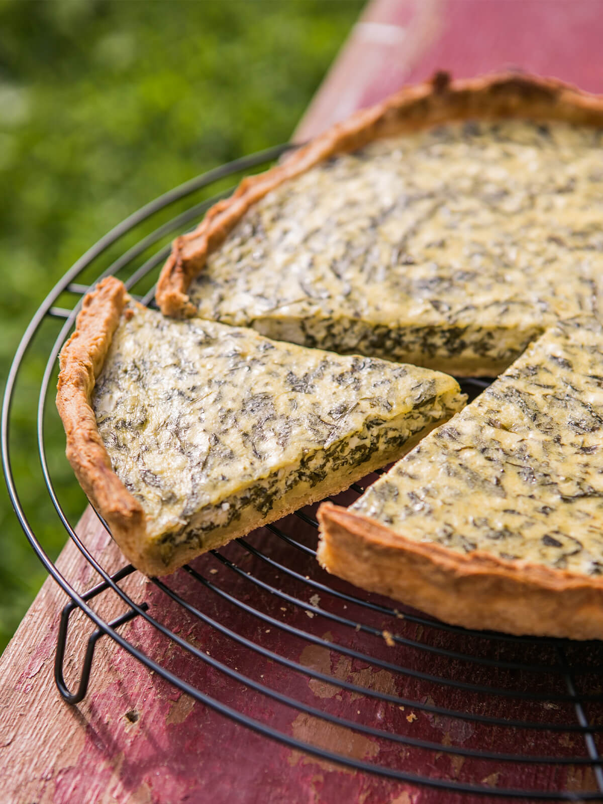 Klassische Spinat-Quiche in Nahaufnahme aus niedrigem Winkel auf einem schwarzen Kuchengitter, ein Stück angeschnitten und leicht abgehoben, mit goldenem Mürbeteigrand und grün gesprenkeltem Eier-Sahne-Guss