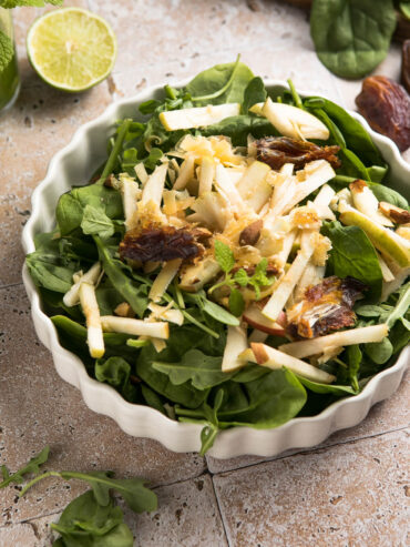 Spinatsalat mit Apfel, Datteln und Mandeln in einer geriffelten cremeweißen Servierform, mit Apfelstiften, weichen Datteln, gerösteten Mandeln und hellem Zitrus-Dressing