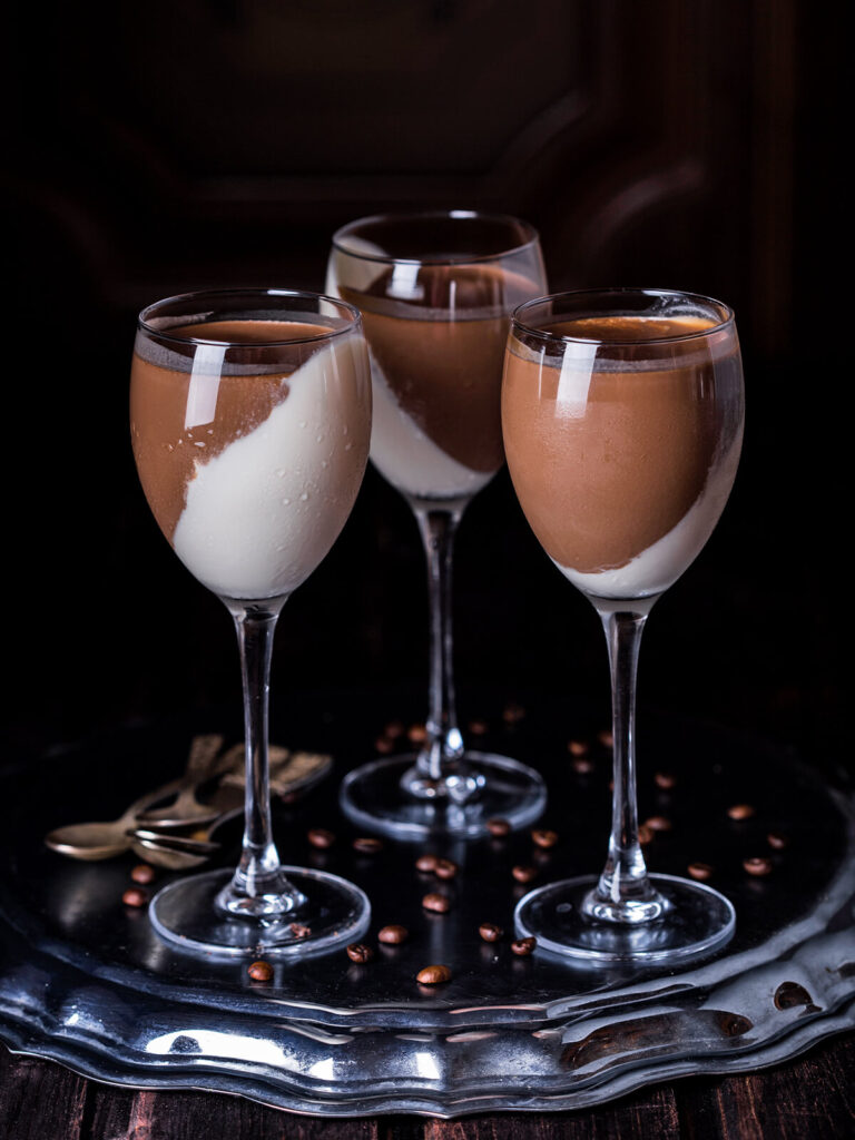 Drei Stielgläser mit zweifarbiger Schoko-Vanille-Panna-Cotta, jede mit diagonaler Trennlinie zwischen weißer und brauner Schicht, auf einer silbernen Platte mit Kaffeebohnen