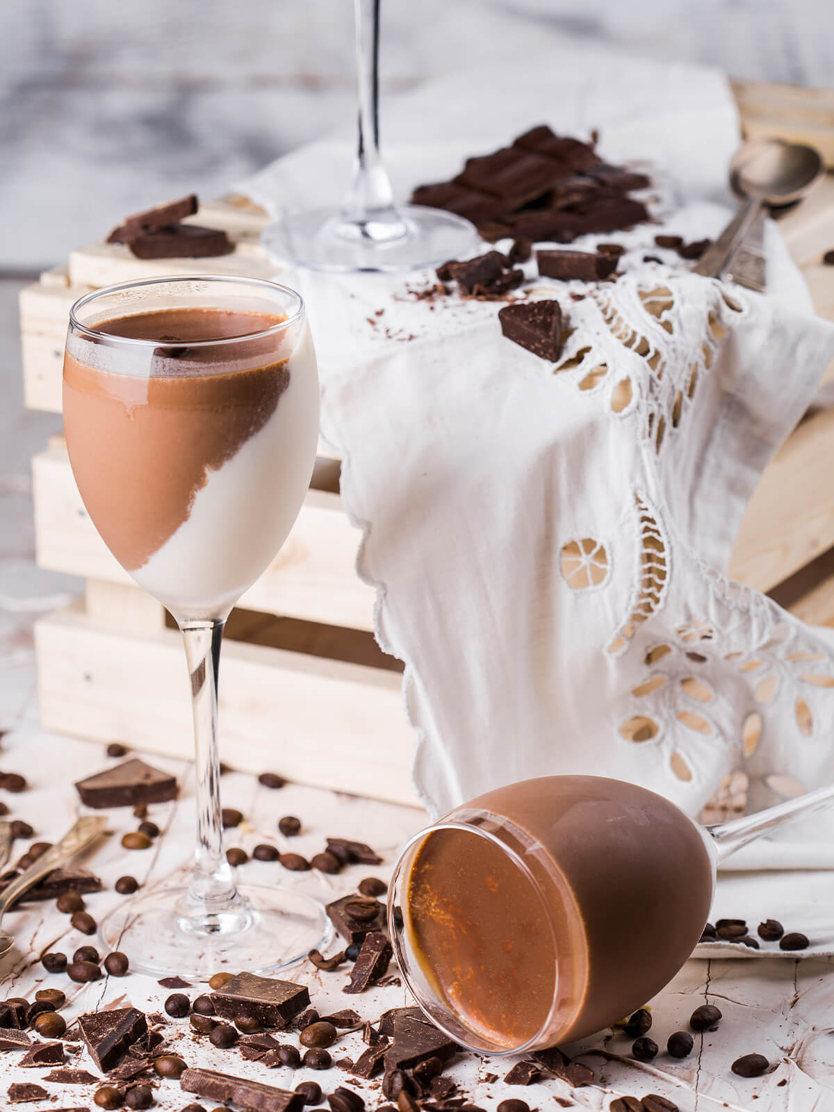 Schoko-Vanille-Panna-Cotta im Stielglas mit diagonaler Trennlinie zwischen weißer Vanille- und brauner Schokoladenschicht, daneben Schokoladenstücke und Kaffeebohnen