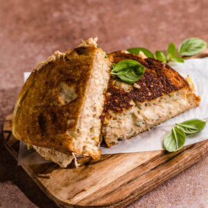 Getoastetes Thunfisch-Sandwich, diagonal halbiert auf einem Holzbrett, mit kräftig bis dunkel gerösteter Kruste, dicker cremiger Thunfischfüllung und frischem Basilikum