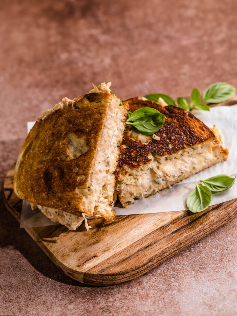 Getoastetes Thunfisch-Sandwich, diagonal halbiert auf einem Holzbrett, mit kräftig bis dunkel gerösteter Kruste, dicker cremiger Thunfischfüllung und frischem Basilikum