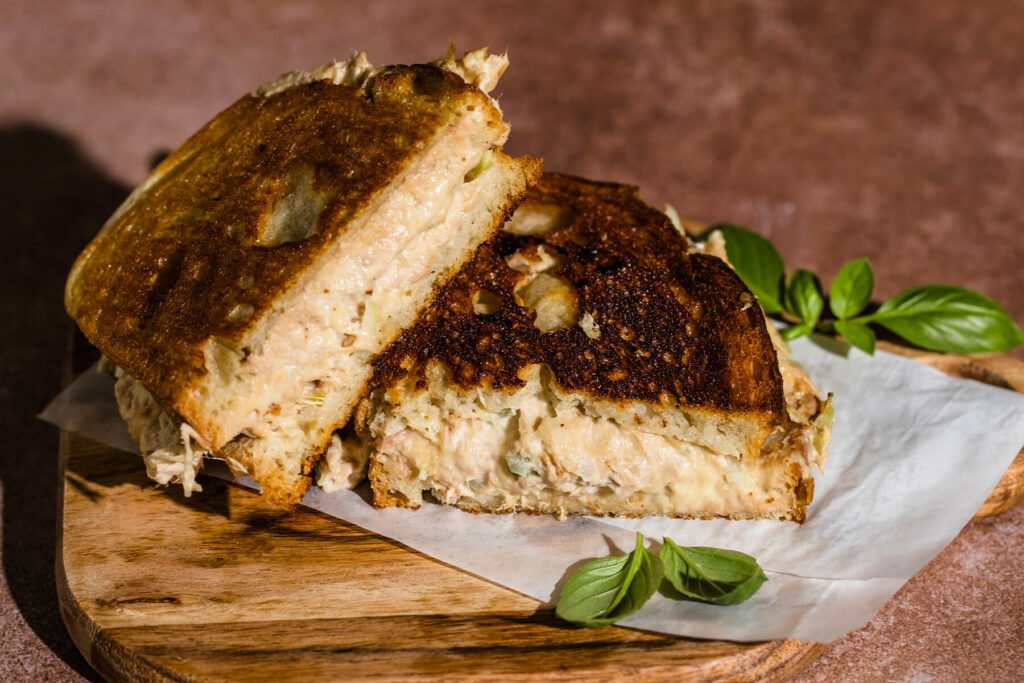 Zwei Hälften des getoasteten Thunfisch-Sandwichs auf Backpapier auf einem Holzbrett, im Anschnitt eine dicke Schicht cremiger Thunfischfüllung mit hellgrünen Selleriewürfeln, die Kruste kräftig bis dunkelbraun geröstet