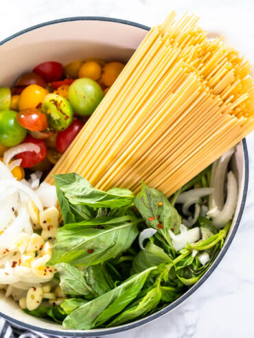 Rohe Zutaten für vegetarische One-Pot-Pasta im Topf – Spaghetti, bunte Kirschtomaten, Zwiebelringe, Knoblauch und frischer Basilikum