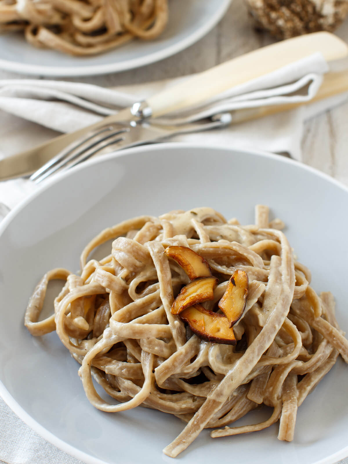 Vollkorn-Tagliolini mit gebratenen Steinpilzen in cremiger Sauce auf weißem Teller, dahinter ein zweiter Teller und ein ganzer Steinpilz