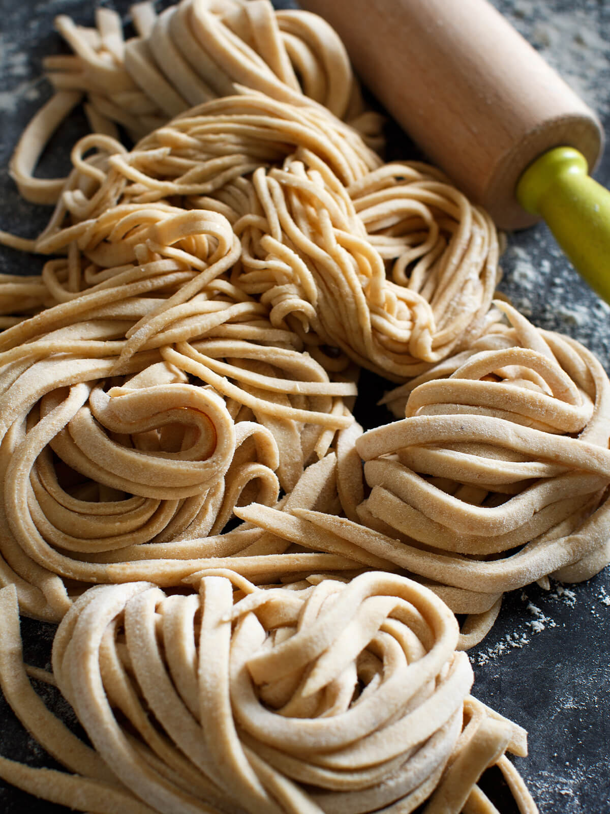 Frische, hausgemachte Vollkorn-Tagliolini als Nester auf dunkler Arbeitsfläche neben einem Nudelholz