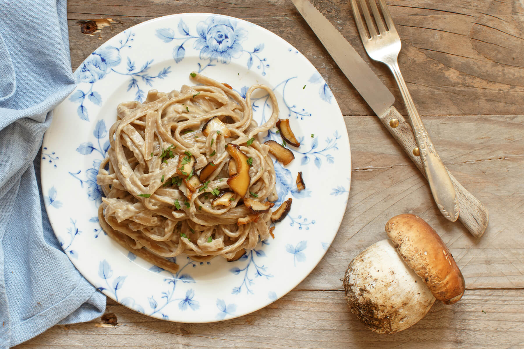 Vollkorn-Tagliolini mit Steinpilzen auf einem Teller mit blauem Blumenmuster, von oben fotografiert, daneben ein ganzer Steinpilz