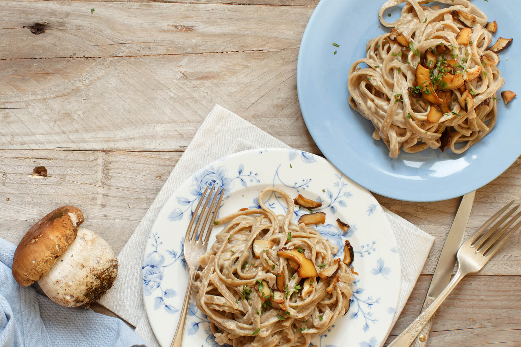 Zwei Teller Vollkorn-Tagliolini mit Steinpilzen von oben, dazu ein frischer Steinpilz und Besteck auf rustikalem Holztisch