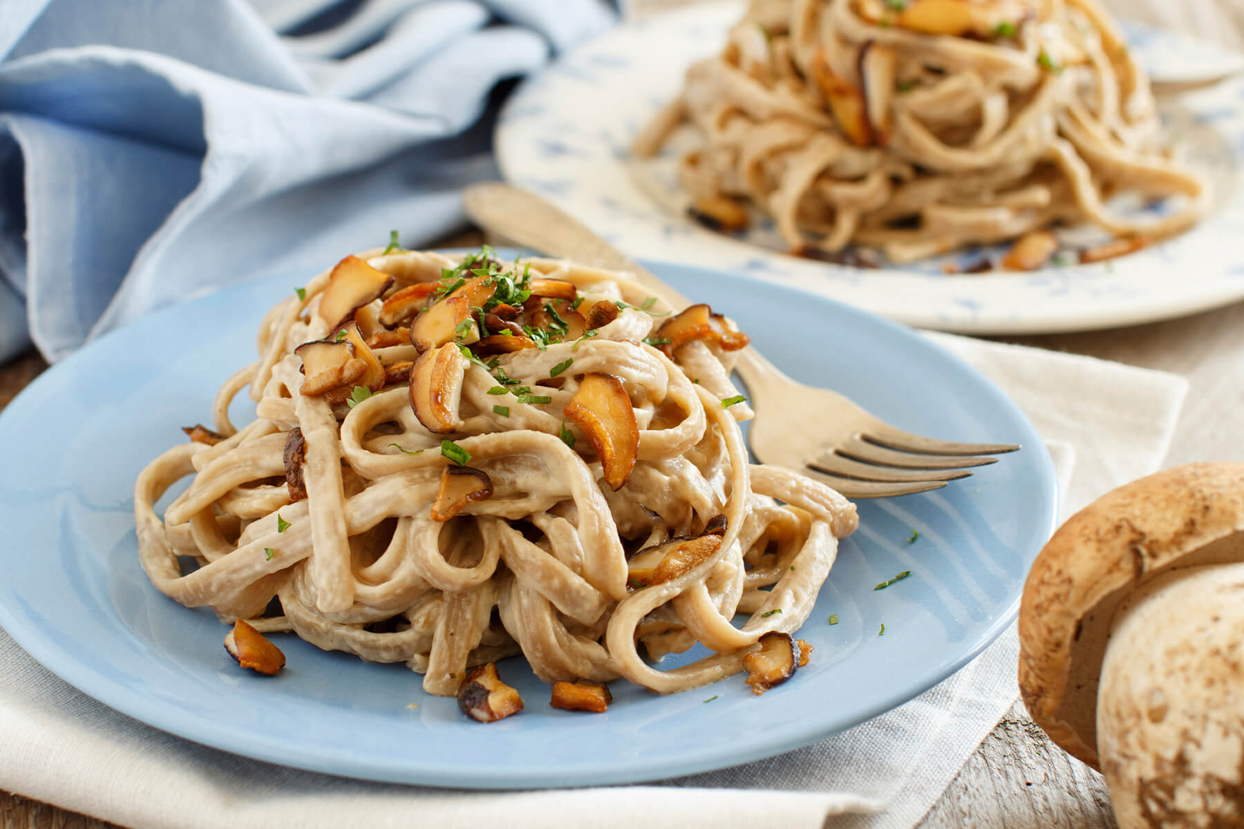 Vollkorn-Tagliolini mit Steinpilzen auf blauem Teller, im Hintergrund ein zweiter Teller und ein frischer Steinpilz
