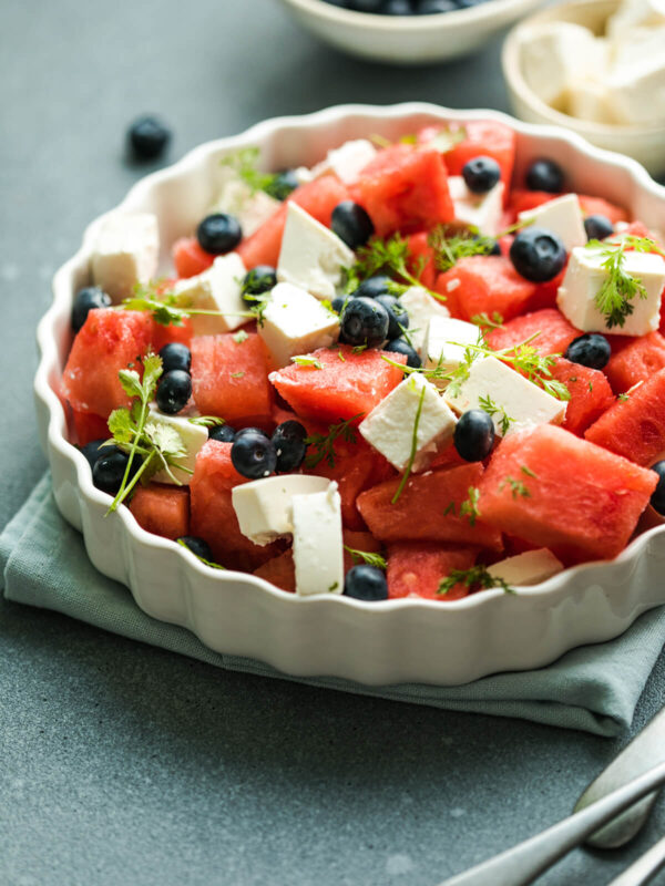 Wassermelone-Feta-Salat mit Heidelbeeren in einer weißen geriffelten Servierform, mit Wassermelonen- und Feta-Würfeln, Heidelbeeren und frischen federartigen Kräutern