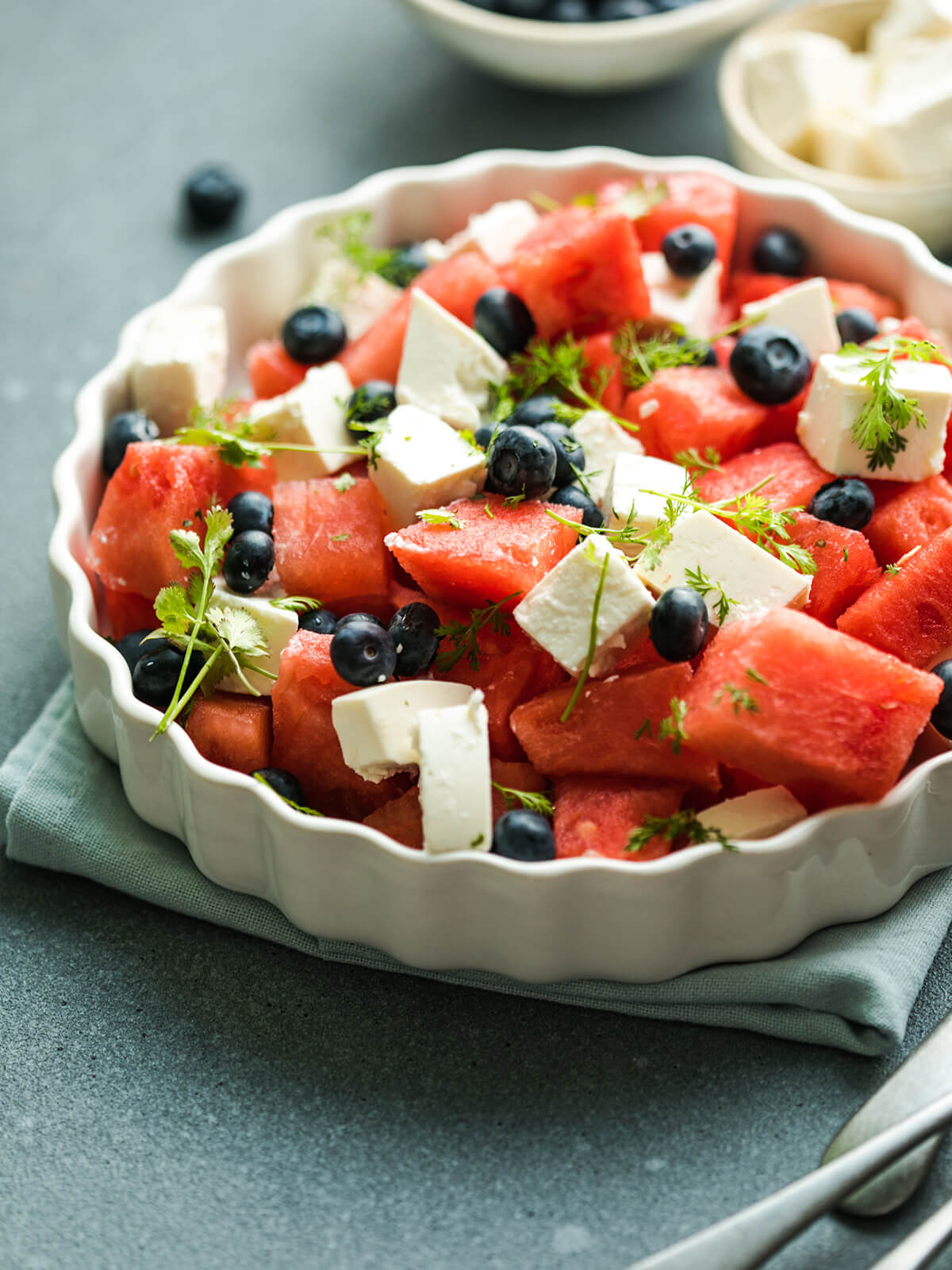 Wassermelone-Feta-Salat mit Heidelbeeren in einer weißen geriffelten Servierform, mit Wassermelonen- und Feta-Würfeln, Heidelbeeren und frischen federartigen Kräutern