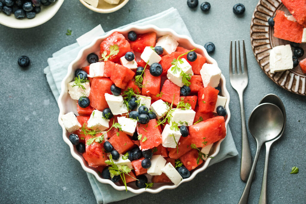 Wassermelone-Feta-Salat mit Heidelbeeren von oben in einer weißen geriffelten Servierform mit frischen Kräutern