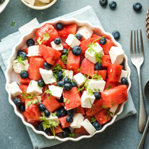 Wassermelone-Feta-Salat mit Heidelbeeren von oben in einer weißen geriffelten Servierform mit frischen Kräutern