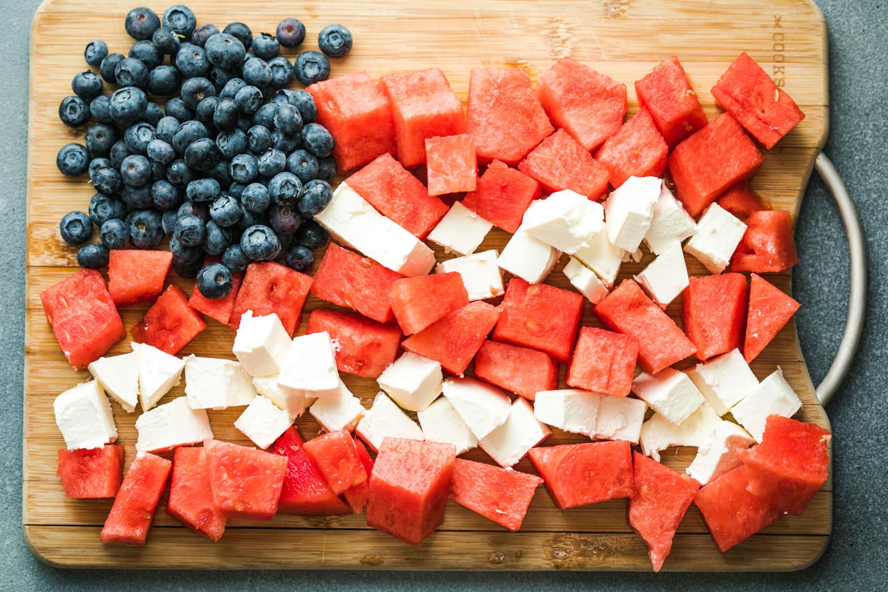 Wassermelonenwürfel, weiße Feta-Würfel und Heidelbeeren auf einem Holzbrett zu einem Flaggenmotiv angeordnet – die Heidelbeeren als blaues Eckfeld, Wassermelone und Feta als rot-weiße Streifen