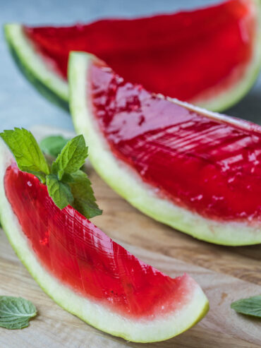 Wassermelonen-Gelee im Melonen-Schiffchen mit Minze – rubinrote, glänzende Gelee-Spalte in grüner Wassermelonenschale auf einem Holzbrett