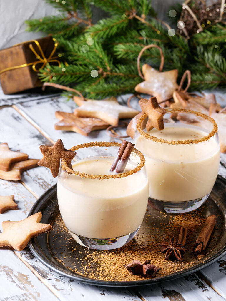 Zwei Gläser cremiger Weihnachts-Eierpunsch mit Zimt-Zucker-Rand, Zimtstange und Zimtstern auf einem Tablett mit Tannenzweigen