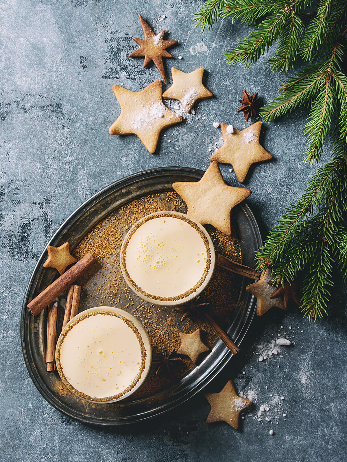 Zwei Gläser Weihnachts-Eierpunsch von oben fotografiert, umrahmt von Zimtstangen, Sternanis, Zimtsternen und Tannenzweigen