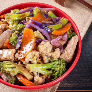 Rote Schale mit Yakisoba-Nudeln, Schweinefleisch und buntem Gemüse aus Brokkoli, Blumenkohl, Rotkohl und Karotte in glänzend-brauner Sauce, daneben Stäbchen