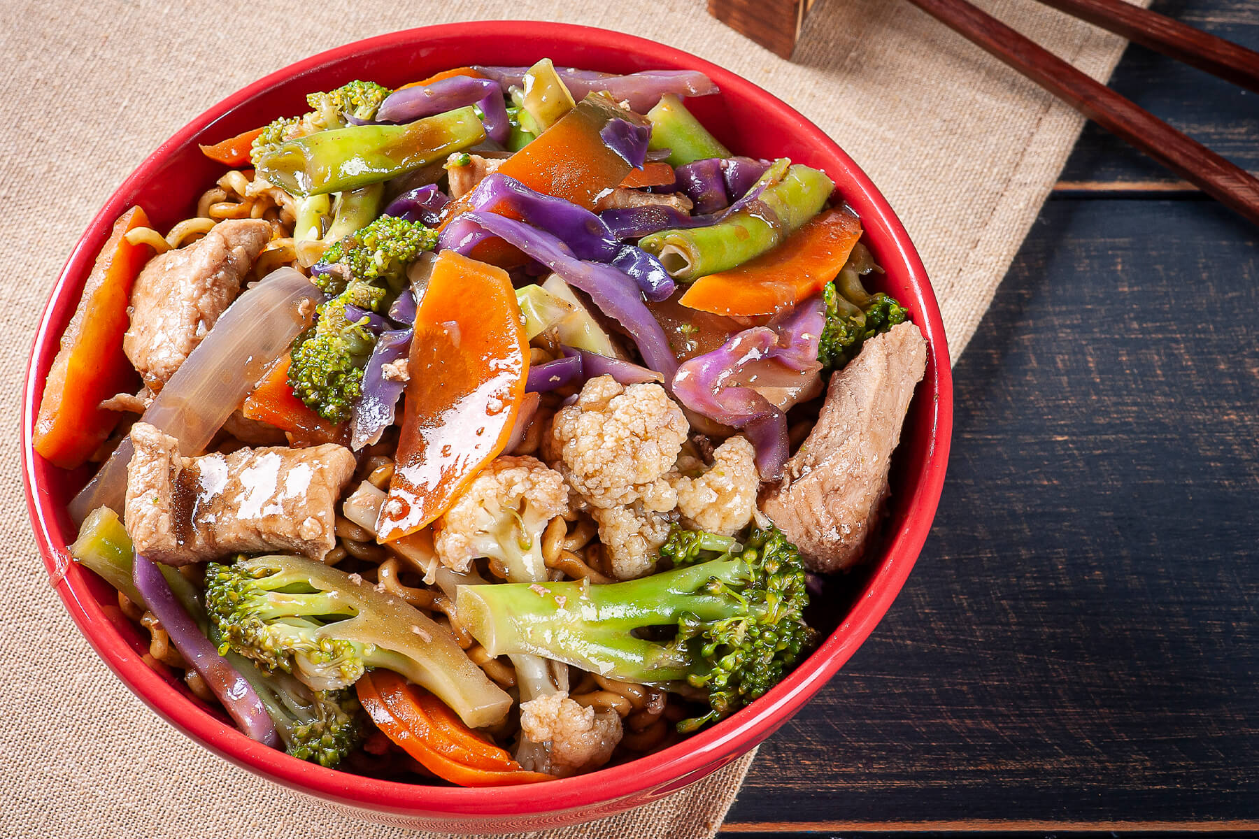 Rote Schale mit Yakisoba-Nudeln, Schweinefleisch, Brokkoli, Blumenkohl, Rotkohl und Karotte in glänzend-brauner Sauce, daneben Stäbchen