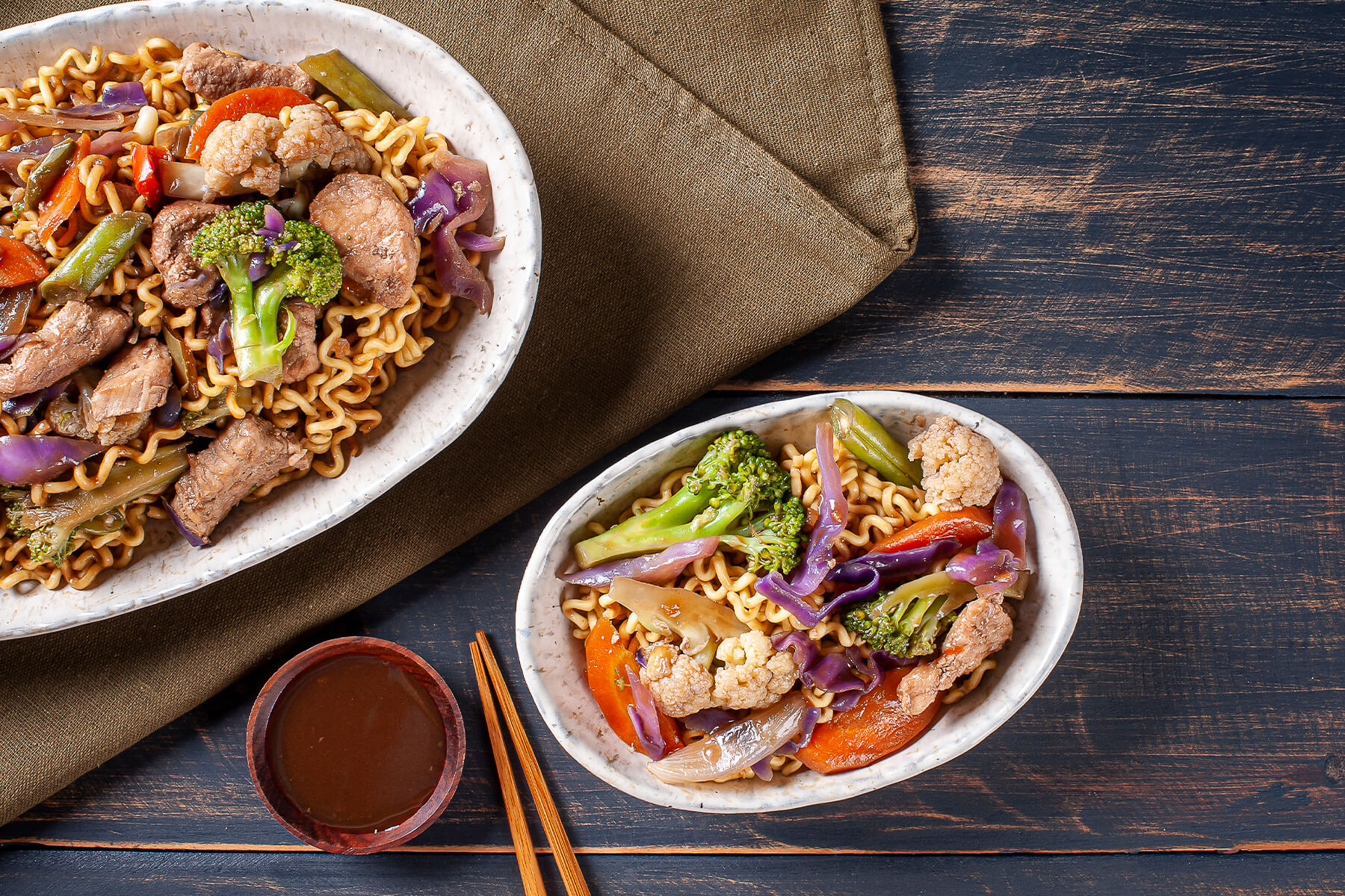 Draufsicht auf eine große und eine kleine Schale Yakisoba mit Schweinefleisch, Brokkoli, Blumenkohl, Rotkohl und Karotte, dazu Stäbchen