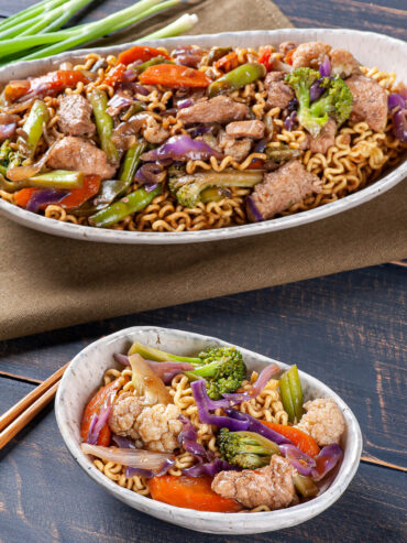 Ovale Schale mit Yakisoba-Nudeln, Schweinefleisch und Gemüse, im Hintergrund eine größere Schale und frische Frühlingszwiebeln