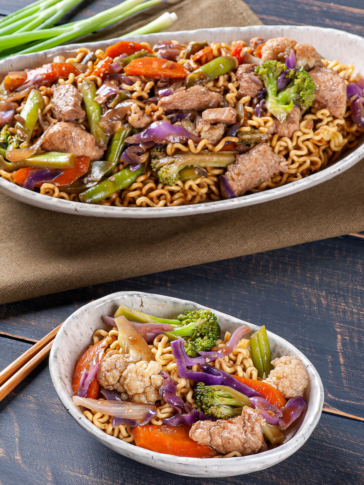 Ovale Schale mit Yakisoba-Nudeln, Schweinefleisch und Gemüse, im Hintergrund eine größere Schale und frische Frühlingszwiebeln