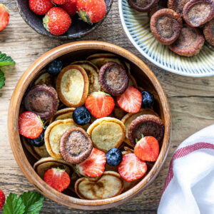 Zweifarbige Mini-Pancake-Cereal von oben: helle Vanille- und dunkle Kakao-Mini-Pfannkuchen gemischt in einer Holzschüssel, dazu Erdbeeren und Heidelbeeren