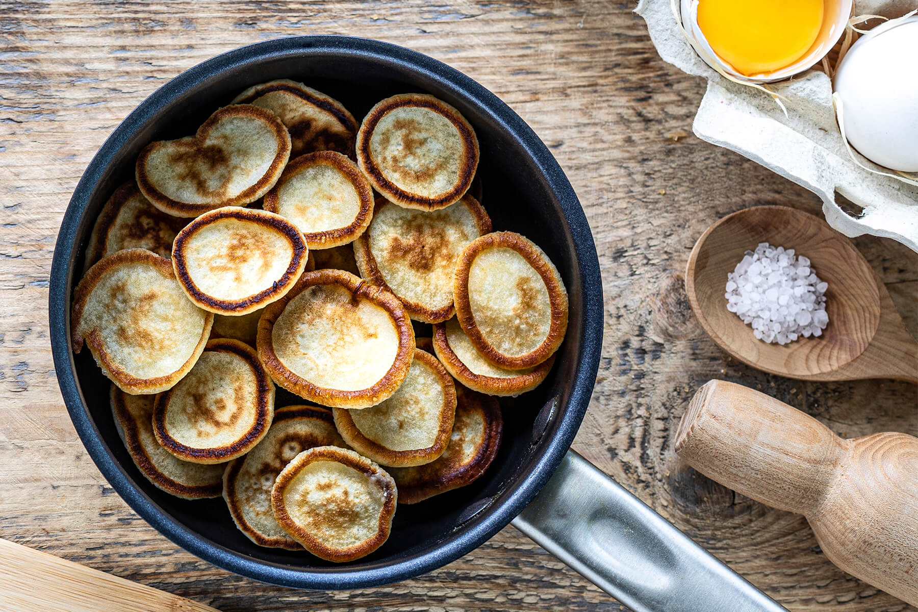 Goldgelbe Vanille-Mini-Pancakes in einer beschichteten Pfanne, daneben Eier und grobes Salz auf einem Holztisch