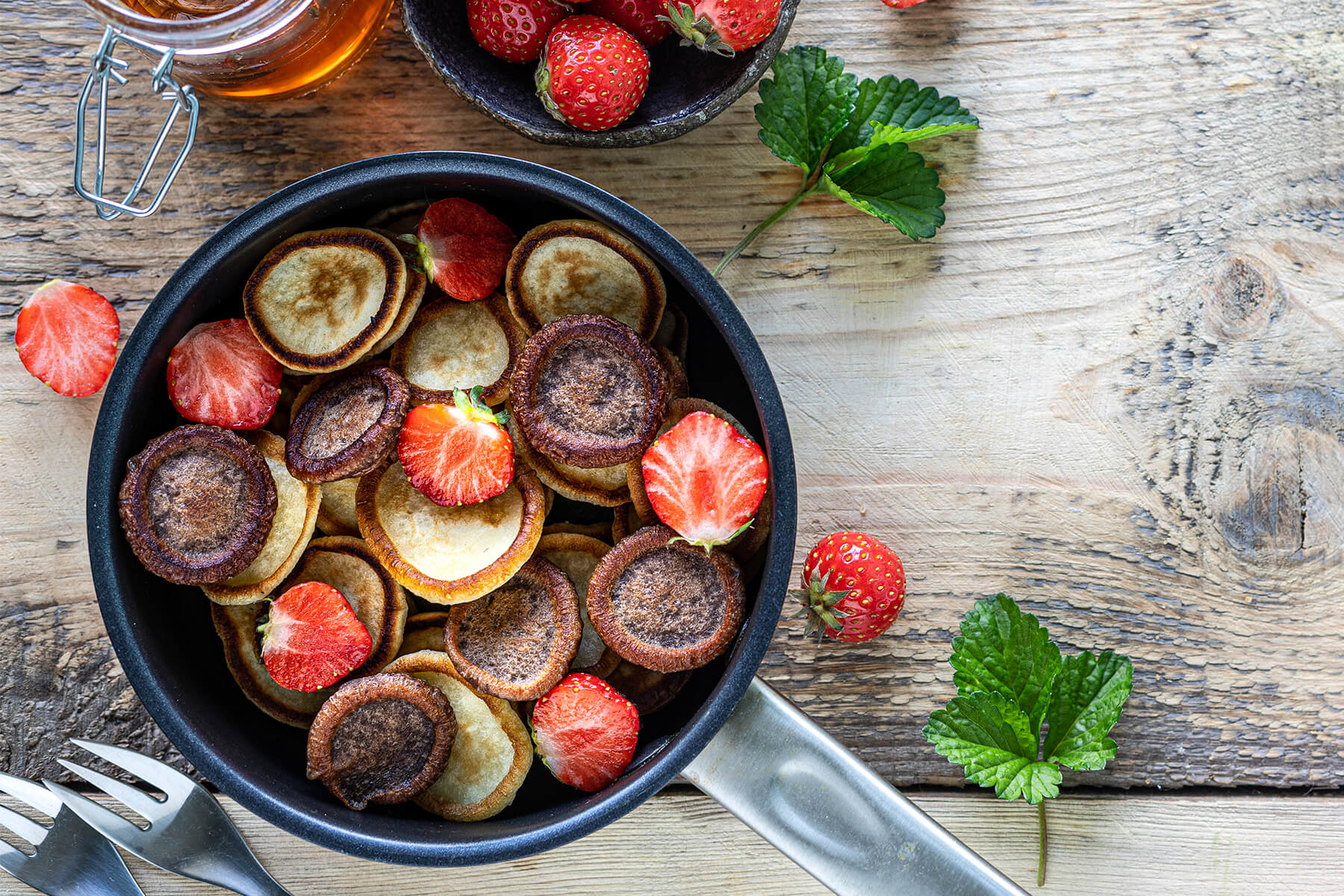 Zweifarbige Mini-Pancakes aus hellem und dunklem Teig in einer Pfanne, daneben Erdbeeren und ein Glas Honig