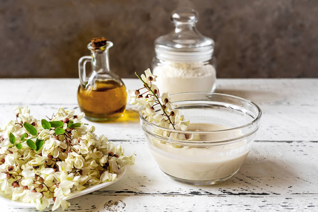 Glasschüssel mit hellem, dickflüssigem Backteig, in die eine weiße Robinien-Blütentraube am Stiel getaucht wird; daneben eine Karaffe mit Öl, ein Glas mit Mehl und ein Teller mit frischen Akazienblüten auf weißem Holztisch