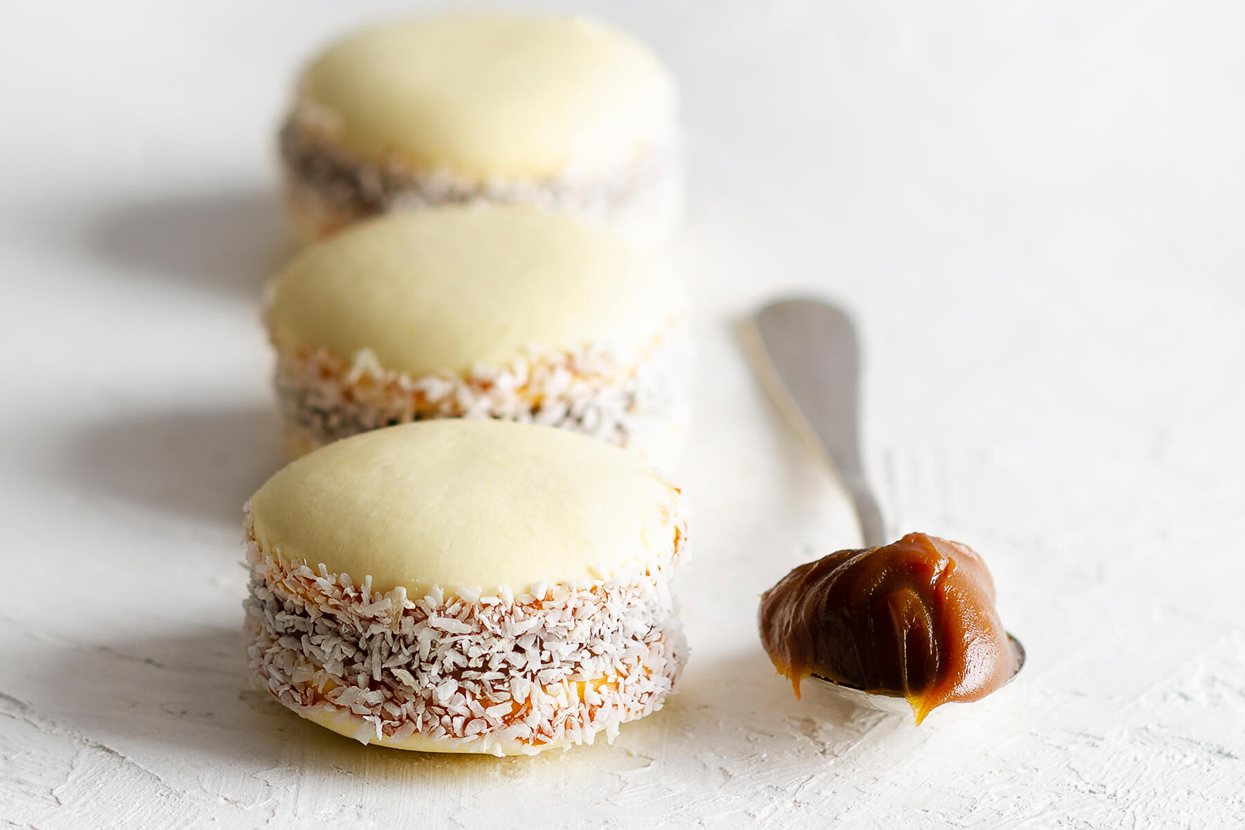 Drei Alfajores de Maicena mit Kokosrand hintereinander, daneben ein Löffel Dulce de Leche
