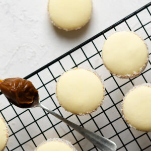 Fertige Alfajores de Maicena mit Kokosrand auf einem schwarzen Kuchengitter, daneben ein Löffel Dulce de Leche