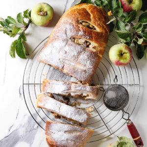 Apfelstrudel aus Hefeteig von oben auf einem Kuchengitter, in Scheiben geschnitten, mit Puderzucker bestäubt, dazu frische Äpfel