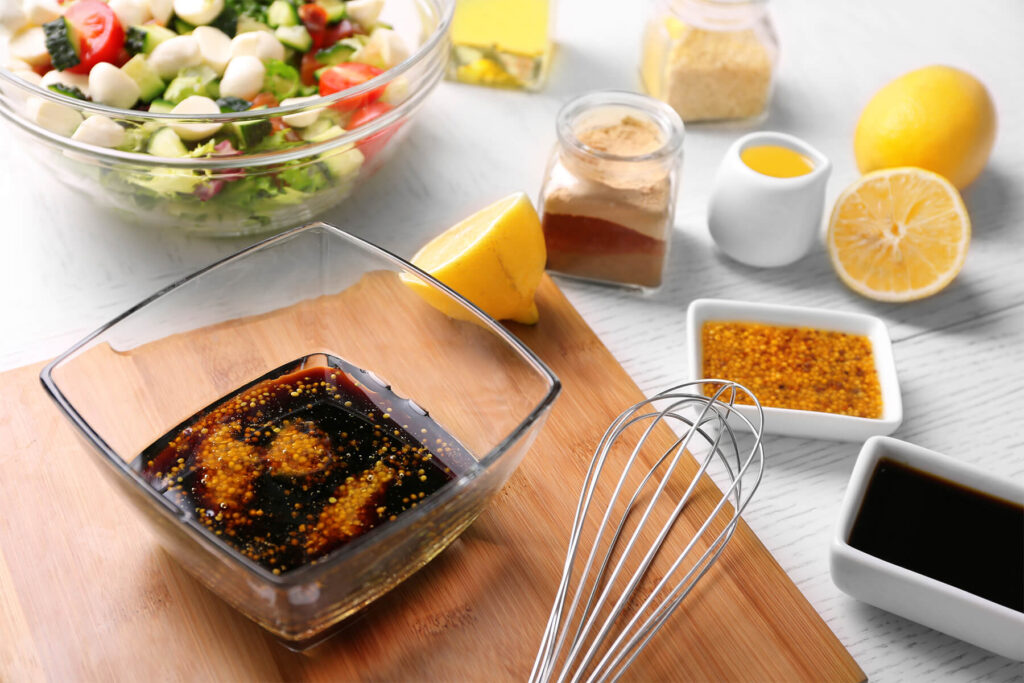 Zutaten für Balsamico-Honig-Senf-Dressing in einer Glasschale: dunkler Balsamico und Körnersenf, daneben Zitrone und ein Schneebesen
