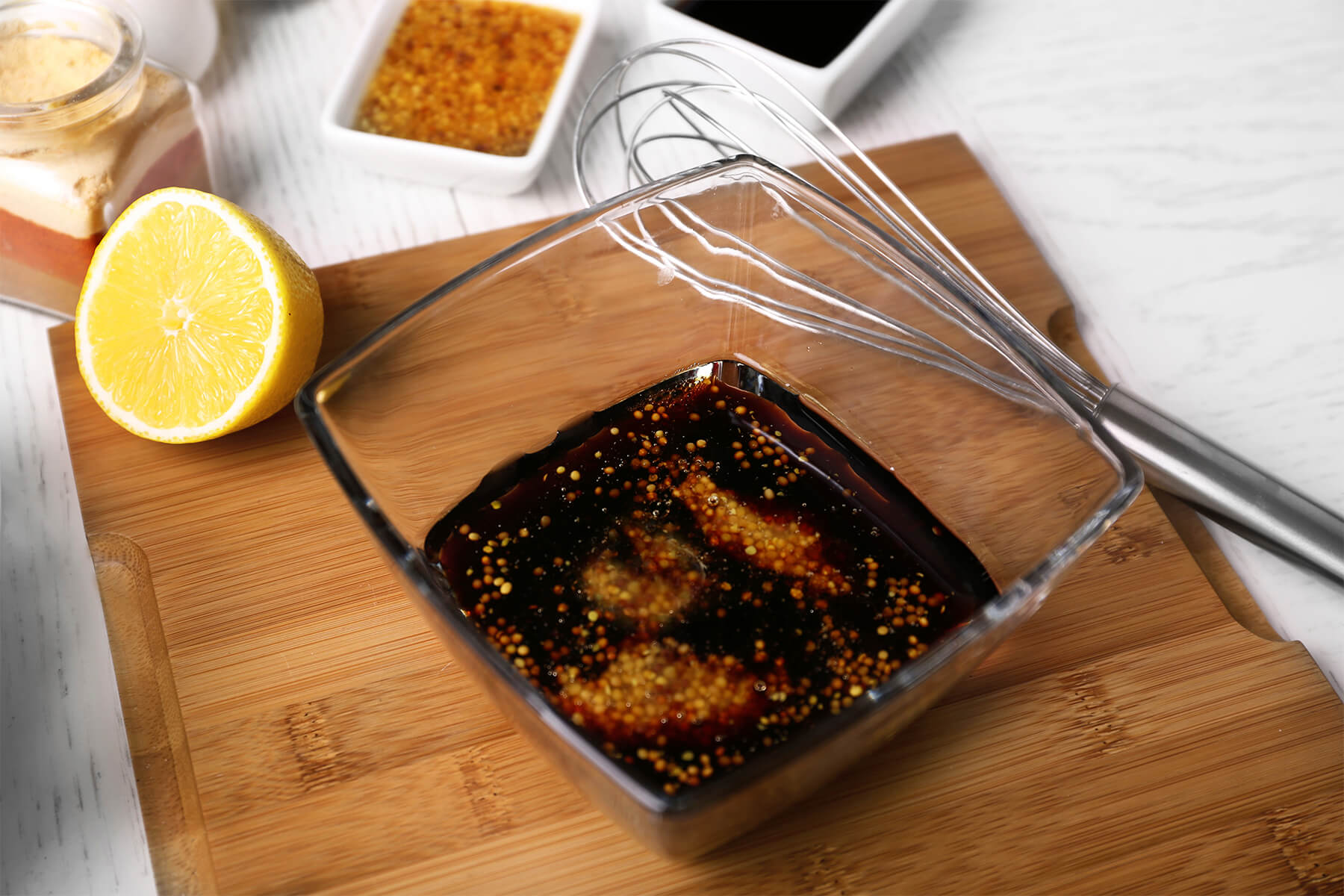 Dunkles Balsamico-Honig-Senf-Dressing mit ganzen Körnersenf-Samen in einer Glasschale, daneben eine halbe Zitrone und ein Schneebesen