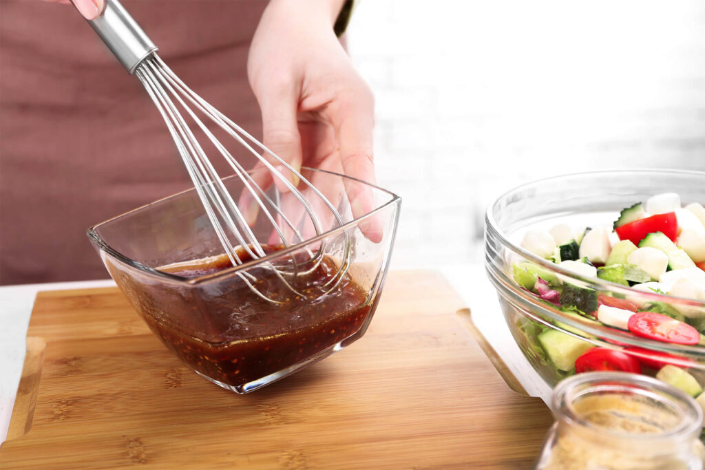 Balsamico-Honig-Senf-Dressing wird mit einem Schneebesen zu einem glänzenden, dunklen Dressing aufgeschlagen, daneben eine Schüssel Salat