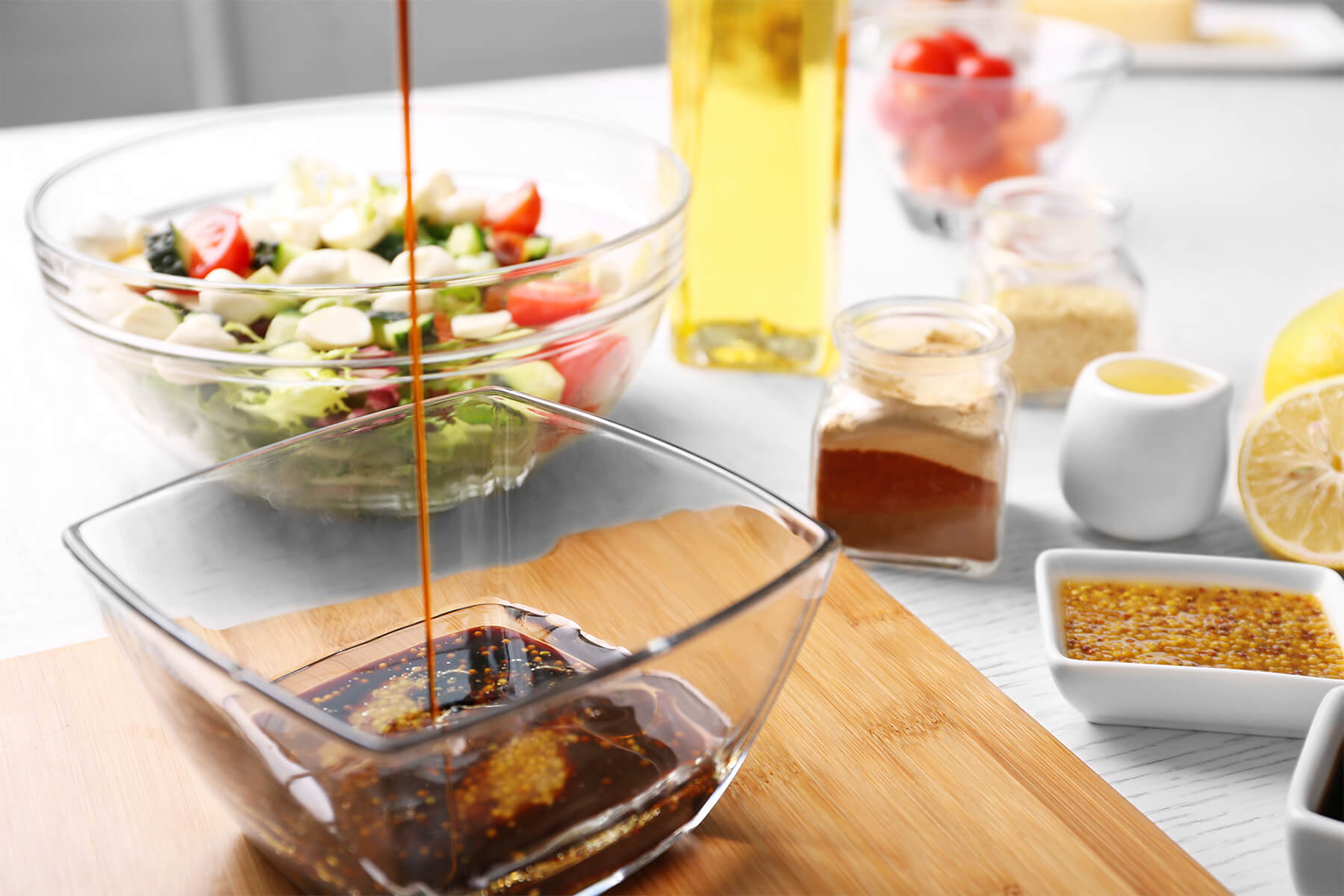 Dunkles Balsamico-Honig-Senf-Dressing wird in eine Glasschale gegossen, im Hintergrund Salat, Olivenöl und Zitrone
