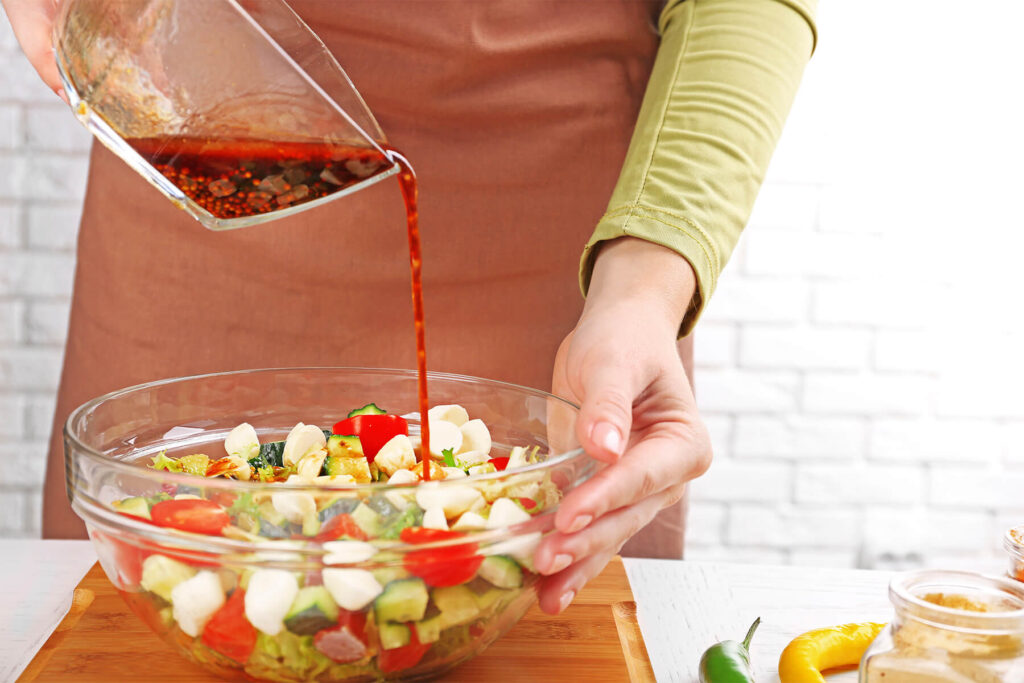 Balsamico-Honig-Senf-Dressing wird aus einer Glasschale über einen Salat aus Mozzarella, Kirschtomaten und Gurke gegossen