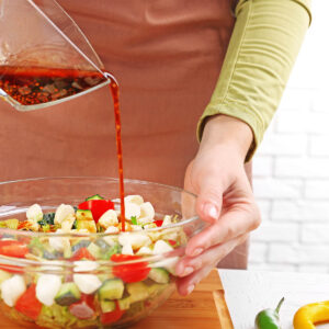 Balsamico-Honig-Senf-Dressing wird aus einer Glasschale über einen Salat aus Mozzarella, Kirschtomaten und Gurke gegossen