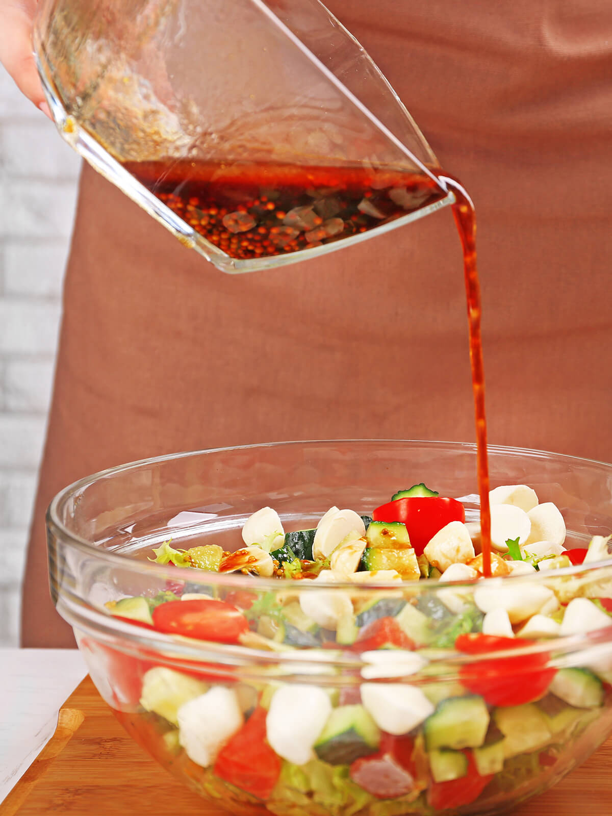 Balsamico-Honig-Senf-Dressing wird aus einer Glasschale über einen frischen Salat mit Mozzarella, Kirschtomaten und Gurke gegossen