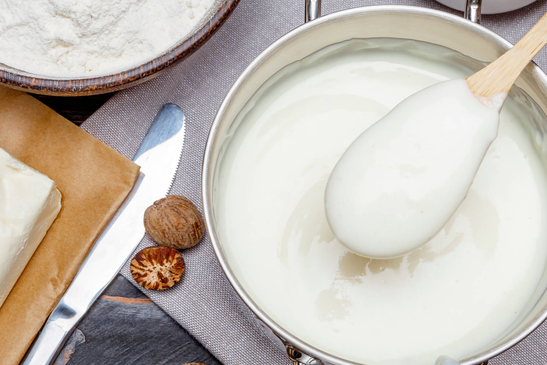 Blick von oben auf cremige Besciamella im Topf mit Löffel, daneben Butter, Mehl und eine Muskatnuss