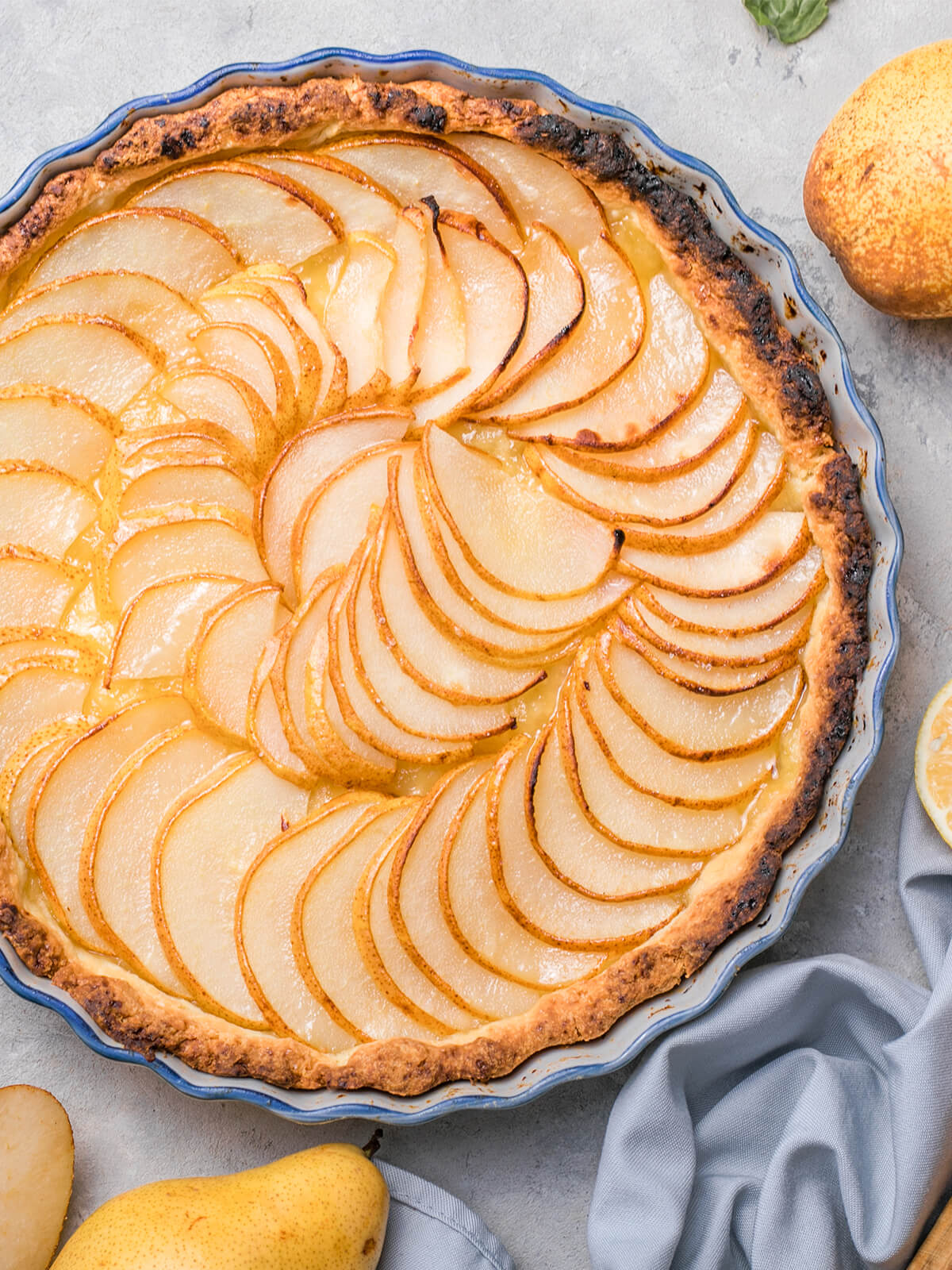 Nahaufnahme einer runden Birnen-Tarte in einer blau geränderten Keramikform mit welligem Rand: hauchdünne, cremefarbene Birnenspalten liegen in zwei ineinandergelegten Kreisen fächerartig überlappend, jede Spalte mit einer feinen goldbraunen Schalenkante voller rostbrauner Pünktchen; dazwischen schimmert eine hellgelbe, glänzende Guss-Schicht, der Teigrand ist kräftig gebräunt und an mehreren Stellen fast dunkel; im Hintergrund heller Steinuntergrund, ein graublaues Tuch, zwei ganze gelbe Birnen am oberen und unteren Bildrand, ein aufgeschnittenes Birnenstück und eine halbe Zitrone