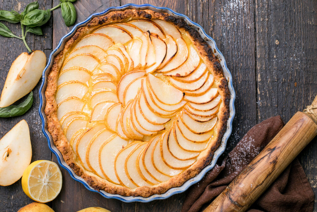 Runde Birnen-Tarte in einer blau geränderten Keramikform mit welligem Rand, von oben auf dunklem Holztisch fotografiert: hauchdünne cremefarbene Birnenspalten fächerartig in zwei ineinandergelegten Kreisen gelegt, jede Spalte mit feiner goldbrauner Schalenkante, dazwischen glänzender hellgelber Guss; der Teigrand ist kräftig gebräunt bis stellenweise dunkel; links liegen aufgeschnittene Birnenstücke mit hellem Fruchtfleisch, eine halbierte Zitrone und frische Basilikumblätter, rechts ein braunes Tuch und ein Nudelholz aus hellem Holz