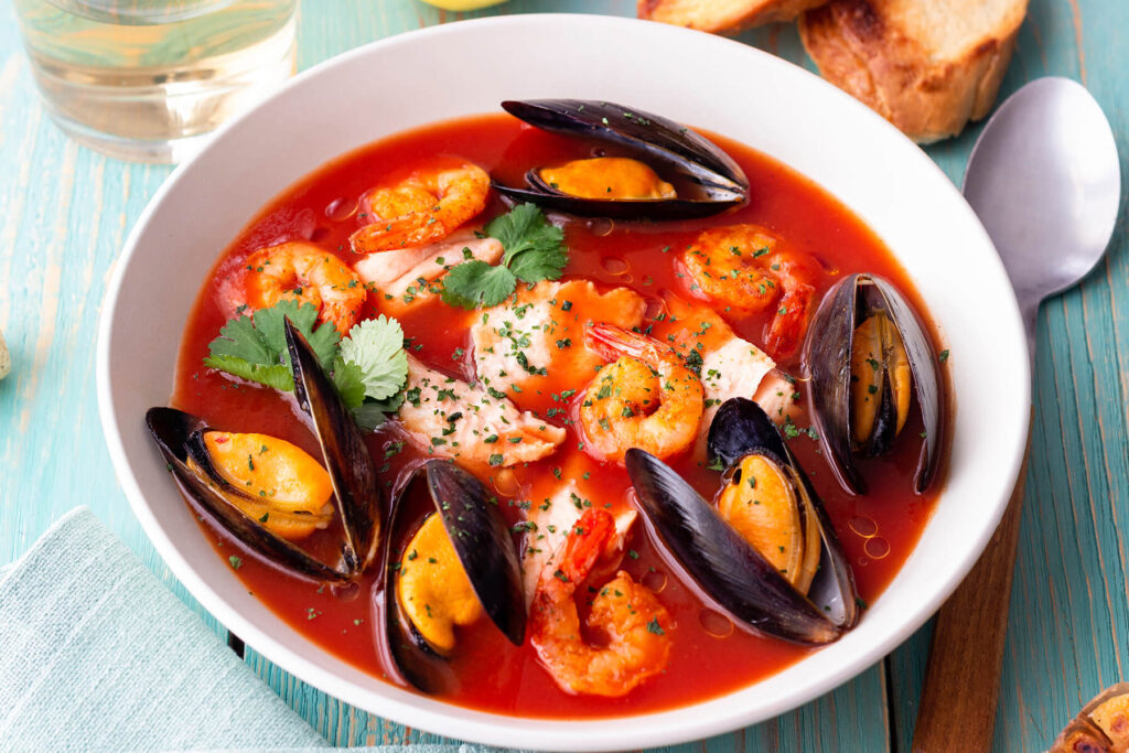 Französische Bouillabaisse mit Fisch, Miesmuscheln und Garnelen in roter Brühe in einem weißen Teller, dazu Baguette