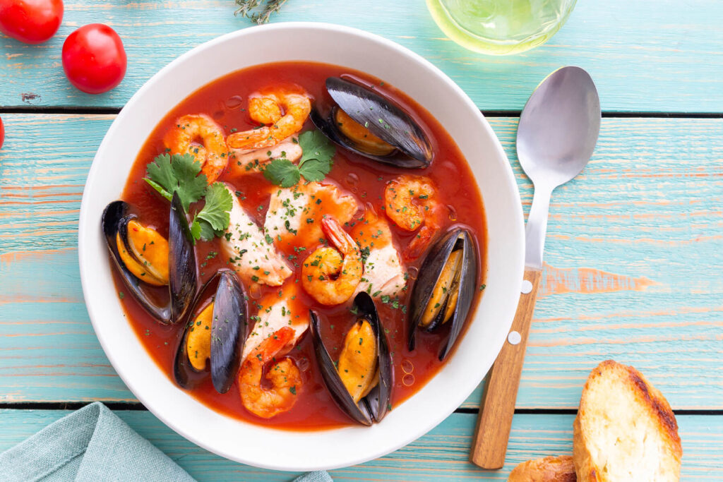 Bouillabaisse von oben mit Garnelen, Miesmuscheln und Fisch, garniert mit Korianderblättern, daneben ein Löffel und Baguette