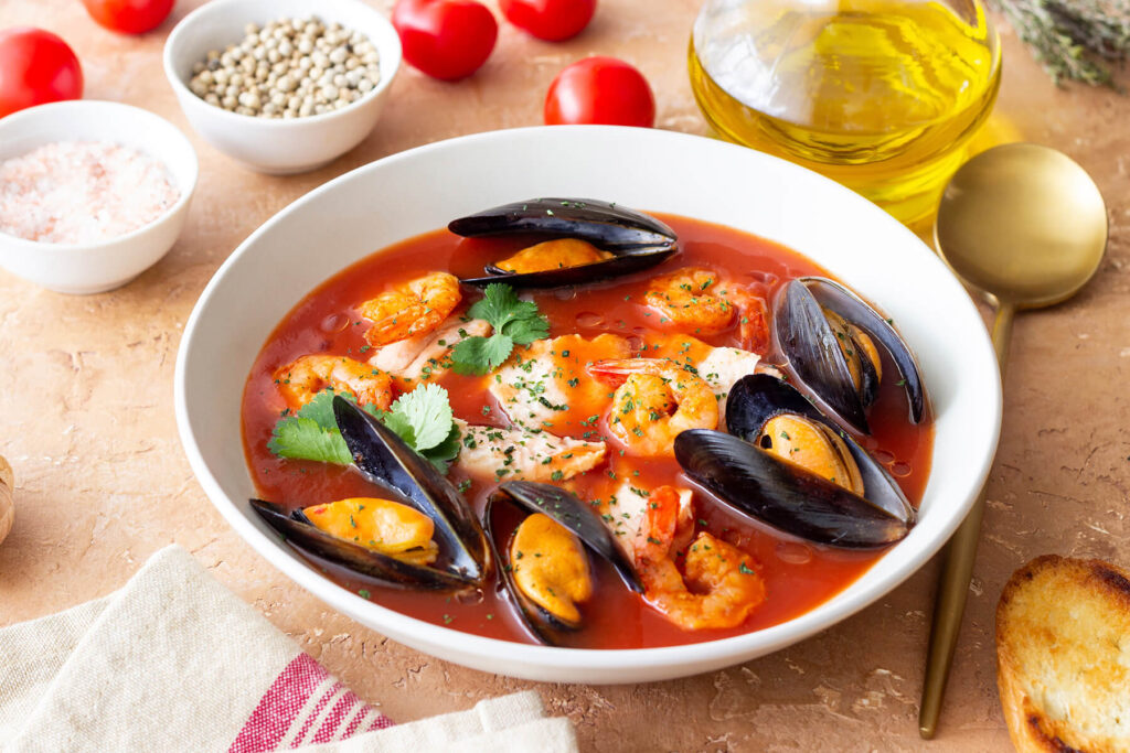 Bouillabaisse in einem weißen Teller mit Miesmuscheln, Garnelen und Fisch, daneben Olivenöl, Tomaten und geröstetes Baguette