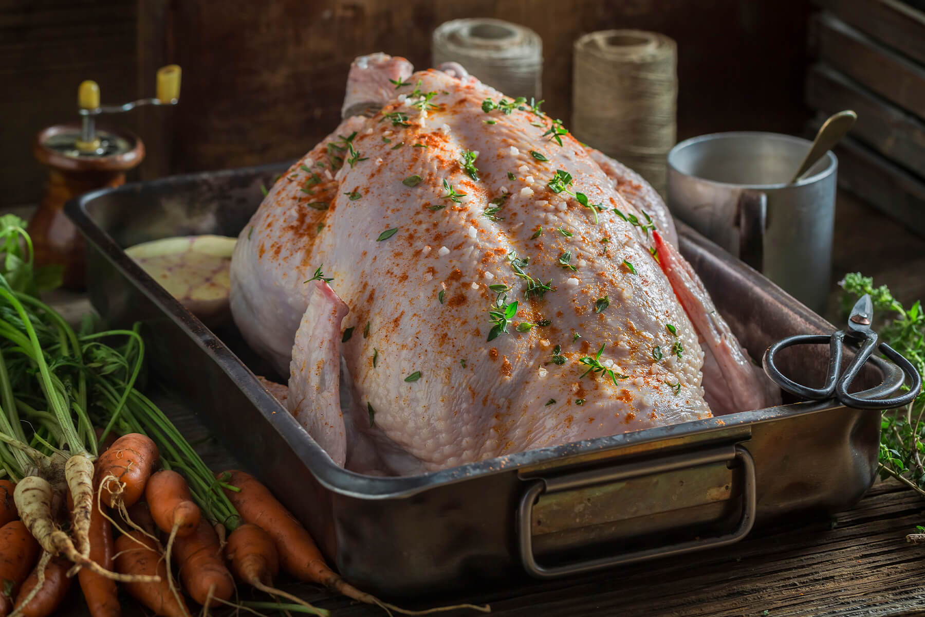 Rohes ganzes Hähnchen mit Salz, edelsüßem Paprika und Thymianblättchen in einer Bratform, daneben junge Karotten und Petersilienwurzeln