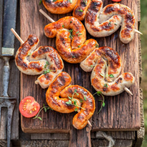 Mehrere gefaltete Bratwürste am Holzspieß auf einem Holzbrett mit halbierten Kirschtomaten und Thymian