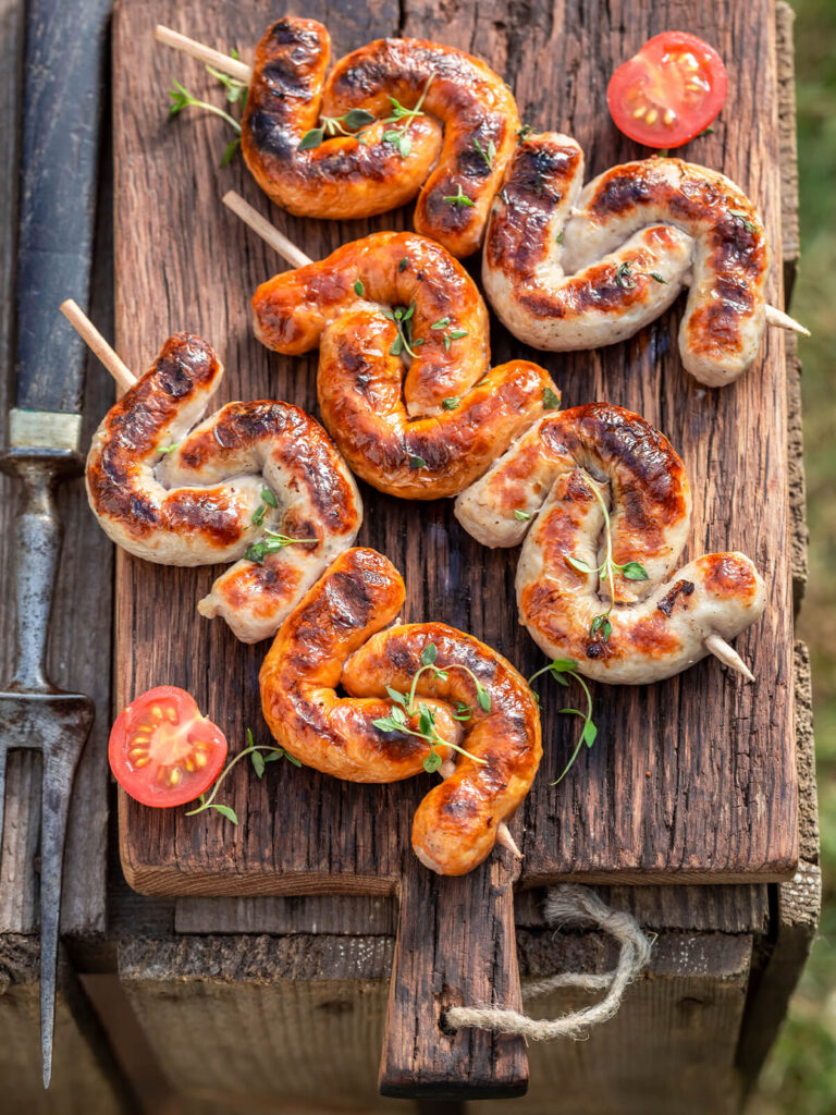 Mehrere gefaltete Bratwürste am Holzspieß auf einem Holzbrett mit halbierten Kirschtomaten und Thymian